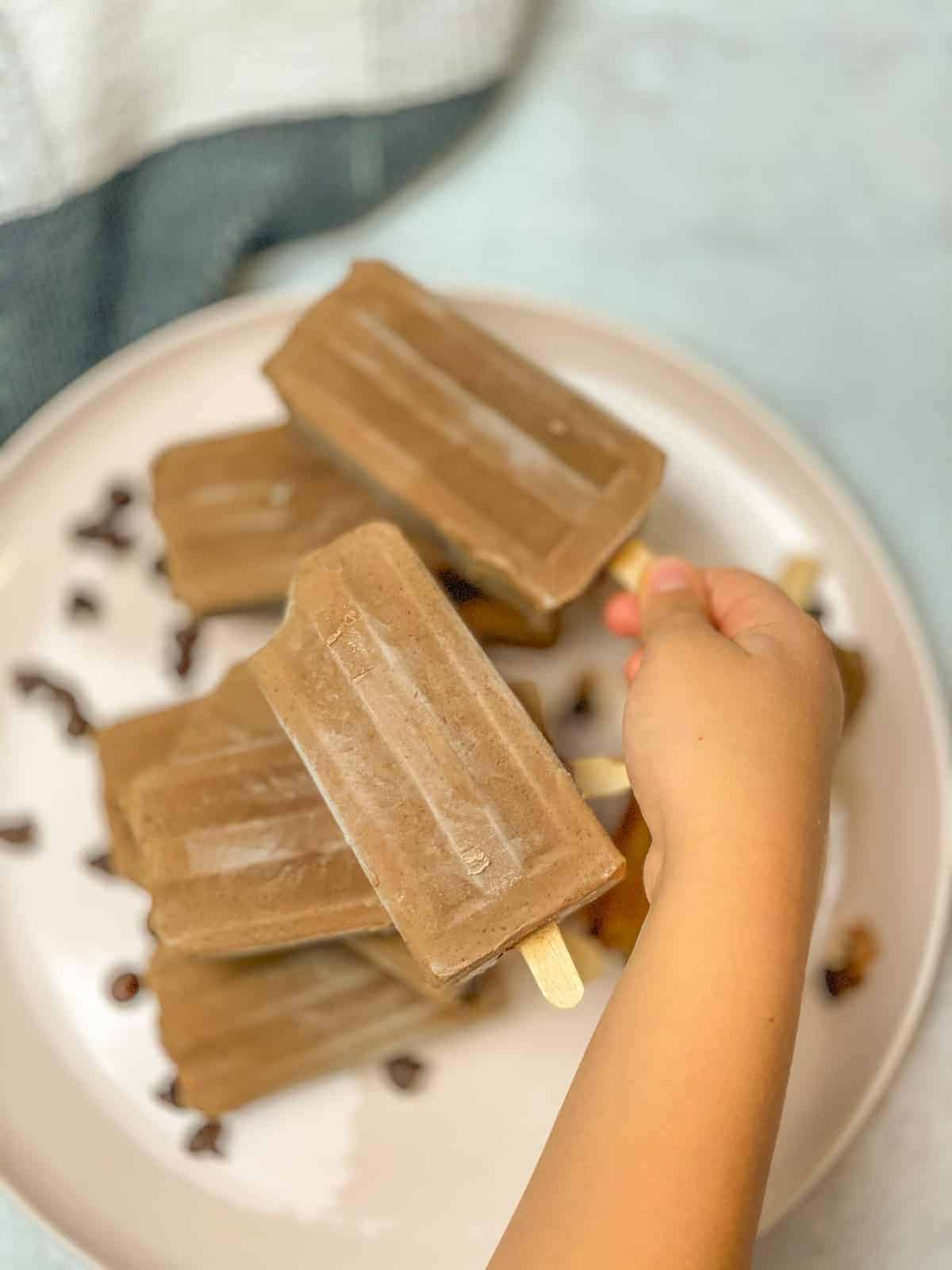 A woman is holding an iced chocolate fudgesicle and there is a dish full of other fudgesicles.