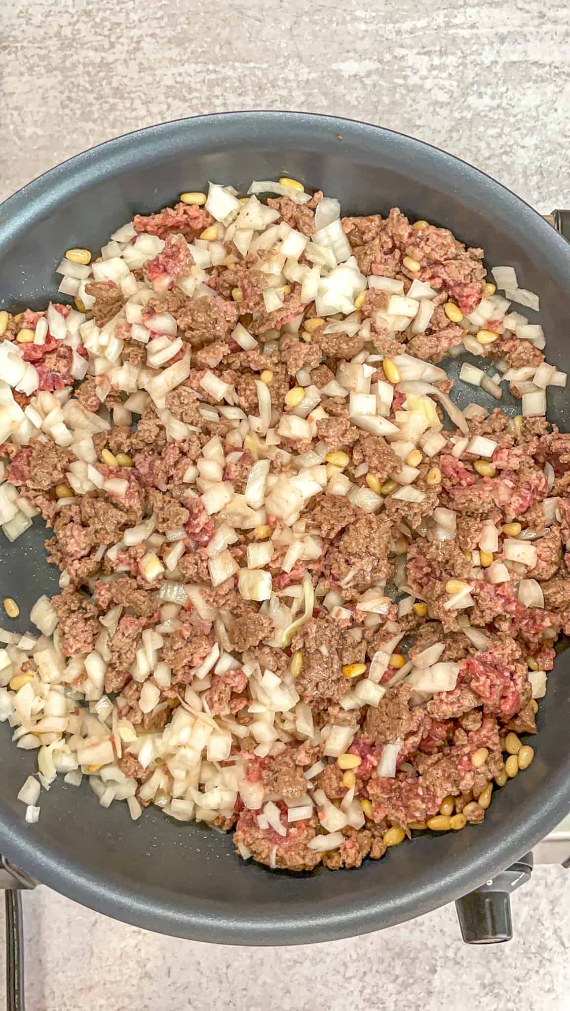 a pan contains translucent onions, pine nuts, and ground beef to be cooked to make the filling of kibbeh bil sanieh