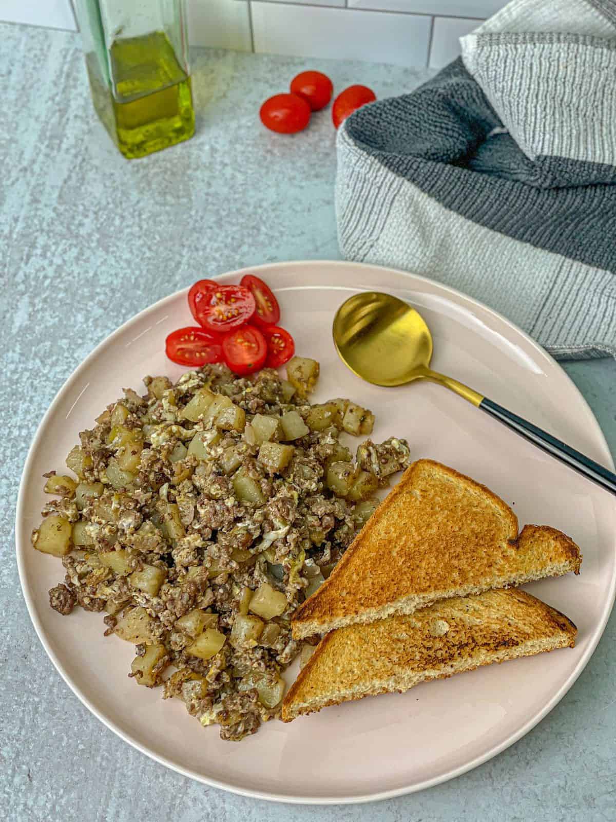 A top view of a large white dish of sautéed spiced beef meat with cubed potatoes, cubed onions, and eggs. It is served with toast bread and halved tomatoes.