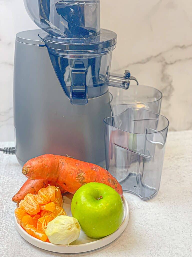 a juicer placed alongside a plate of raw juice ingredients: raw juice made up of sweet potato, mandarin, green apple, and lemon