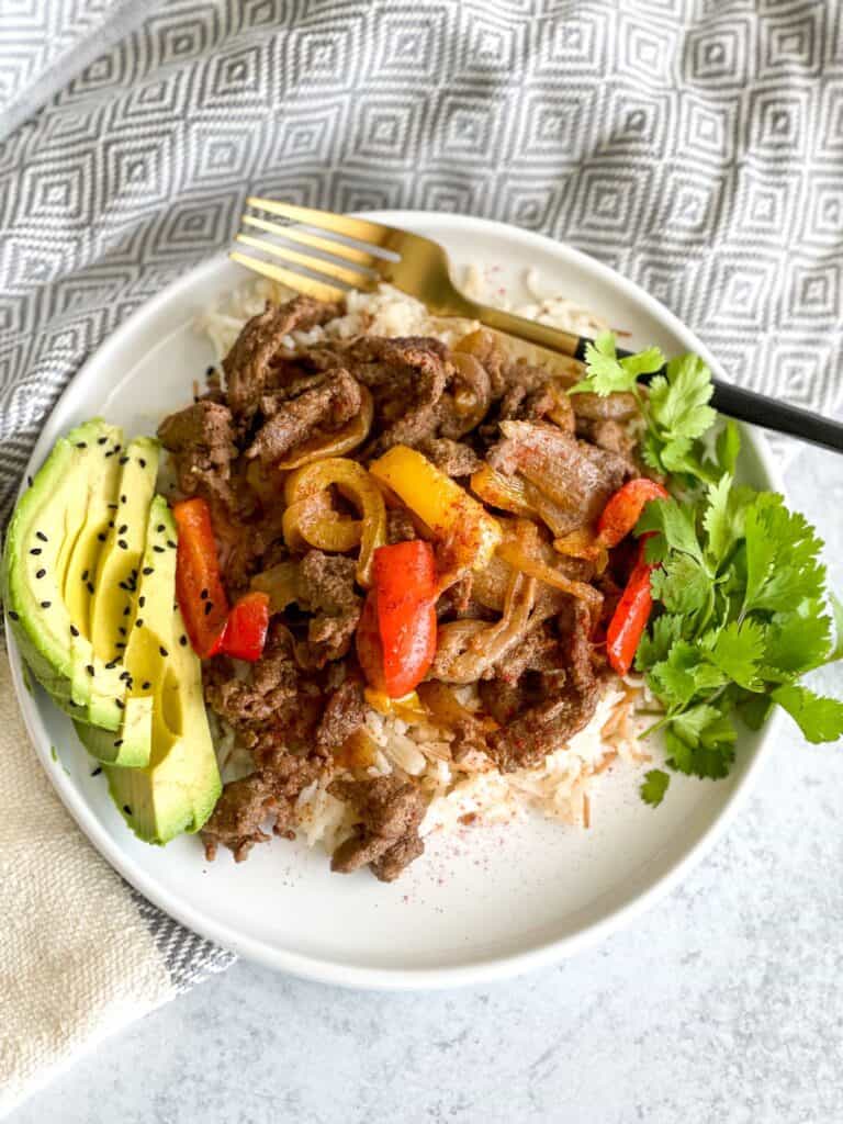 A hearty beef steak fajita bowl with peppers and onions on a bed of white vermicelli rice and a side of avocado