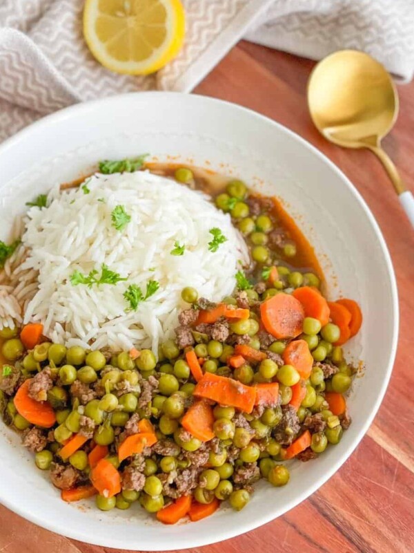 A plate of pea and carrot stew featuring ground beef and a side of white rice with one half of lemon on the side.
