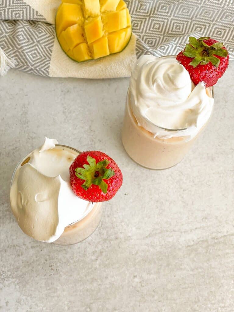 Two glass cups filled with strawberry mango banana smoothie, topped with whipped cream, and decorated with a strawberry. Half a mango fruit is placed beside the cups.