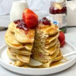 A tall stack of fluffy single-serve pancakes topped with strawberry jam and fresh strawberries, drizzled with syrup, with a bite cut out.