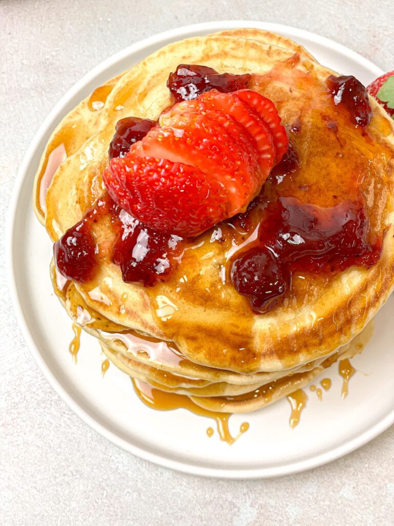 Almond milk pancakes topped with strawberry jam and maple syrup and garnished with fresh strawberries. They are temptingly scrumptious!