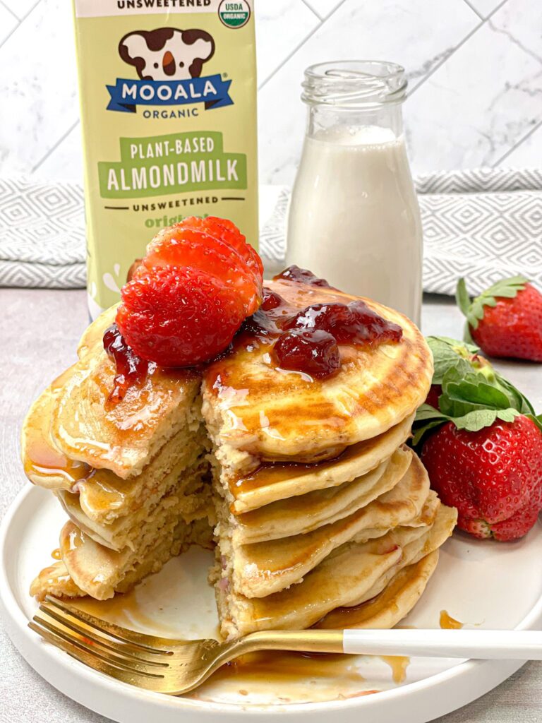 Almond milk pancakes stacked above each other and drizzled with maple syrup and strawberry jam. Fresh strawberries add a kick to this delectable dish.
