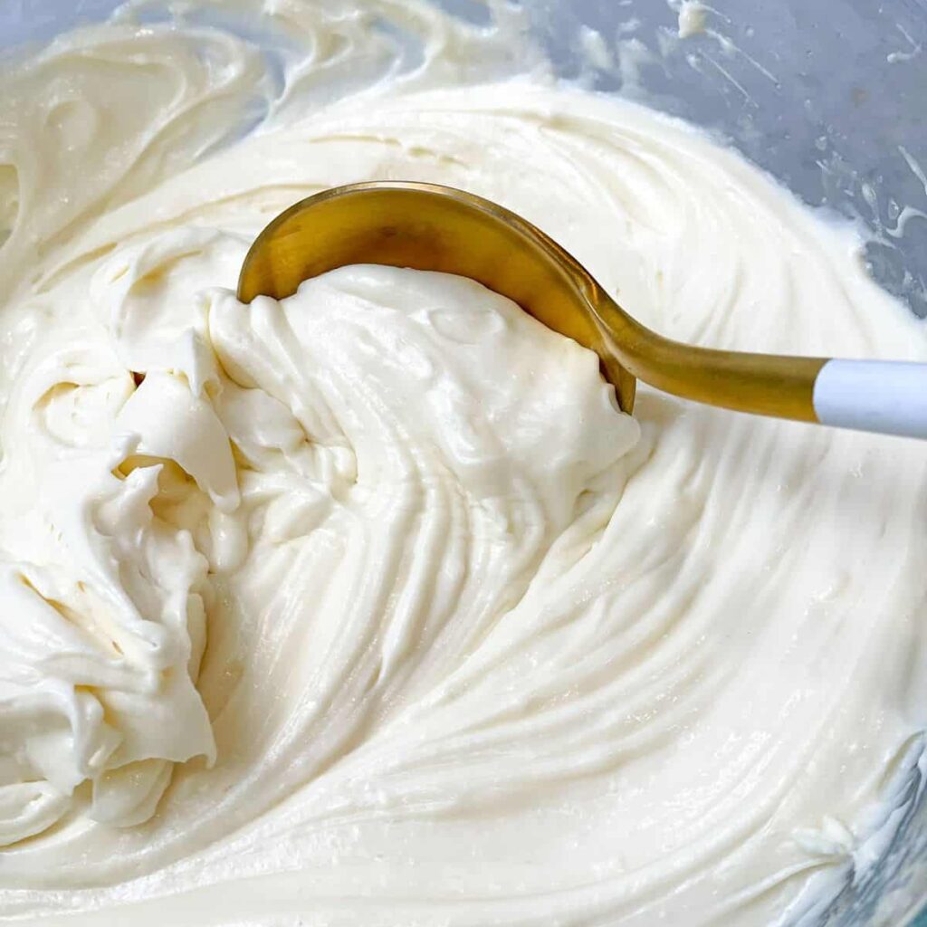 Cream Cheese Frosting Indulgent cream cheese frosting in a glass bowl, with a lavish golden spoon poised for a delightful scoop. The luxurious swirls of the frosting glisten, promising a heavenly blend of sweetness and sophistication in every golden-touched bite.