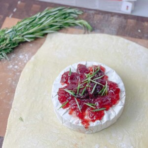 A block of brie cheese topped with red jam and herbs is placed on top of a rolled out puff pastry dough.