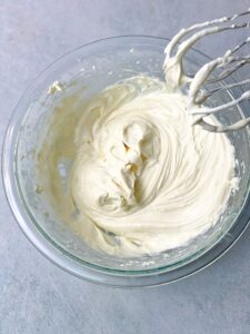 A sumptuous pool of cream cheese frosting fills a glass bowl, enticing with its silky texture and rich aroma.