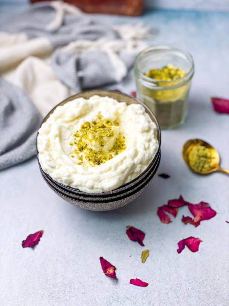 A bowl filled with clotted cream and garnished with some ground pistachios. Some dried rose petals, a spoon filled with ground pistachios, and a ground pistachio jar, decorate the area around the ashta bowl.