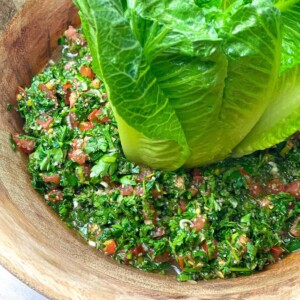 A bowl filled with Taboule Salad that is made up of chopped parsley, tomatoes, onions, and other greens. The bowl is decorated with some lettuce leaves.