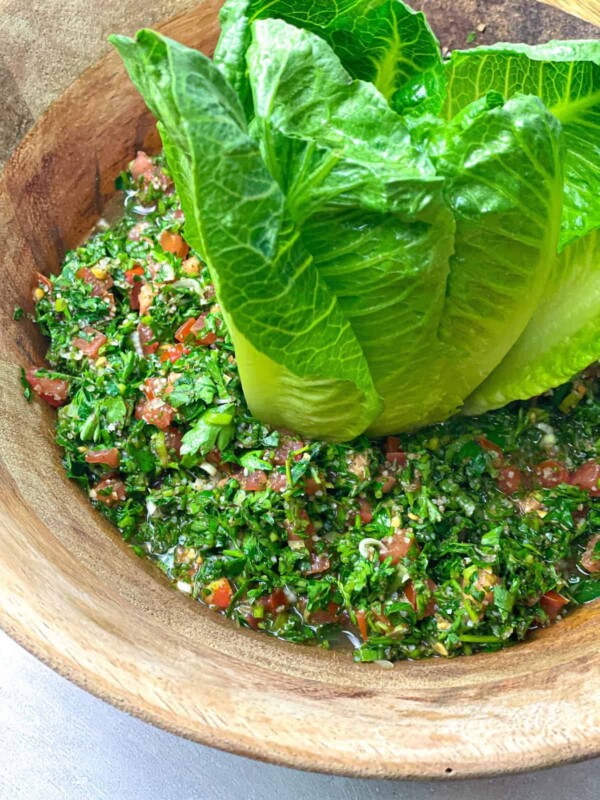 A bowl filled with Taboule Salad that is made up of chopped parsley, tomatoes, onions, and other greens. The bowl is decorated with some lettuce leaves.