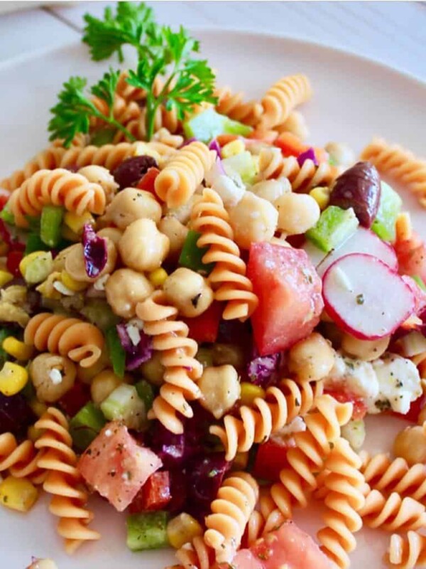 A plate of Greek pasta salad featuring chopped tomatoes, radishes, chickpea, corn, green bell pepper, and fusilli pasta.