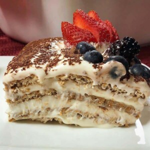 A piece of Nescafé cake featuring layers of biscuits and cream topped with cocoa powder black berries, blue berries, and strawberries.