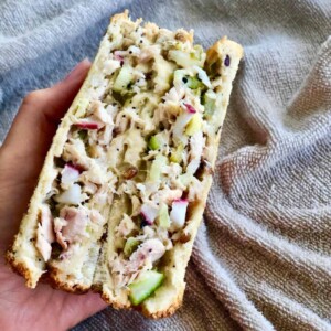 A hand holding a mix of tuna, pickles, radish, gouda cheese and chopped celery sandwiched in toasted bread.