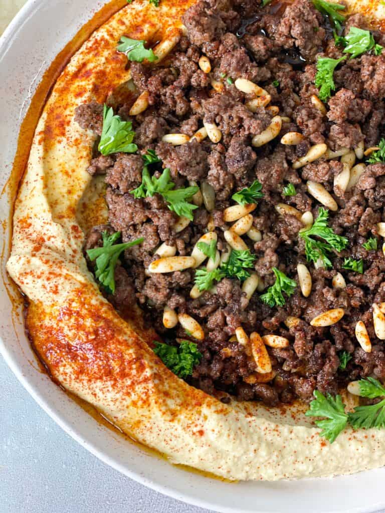 close up of a hummus with spiced meat on top garnished with pine nuts and parsley