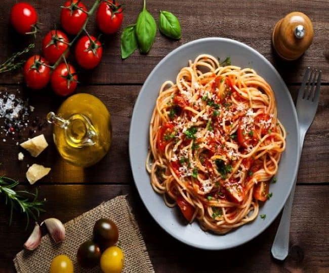 A metal plate of spaghetti with kid sauce is on a table. Around the plate, there are some roma tomatoes, basil leaves, a small jar of olive oil, and a fork. 