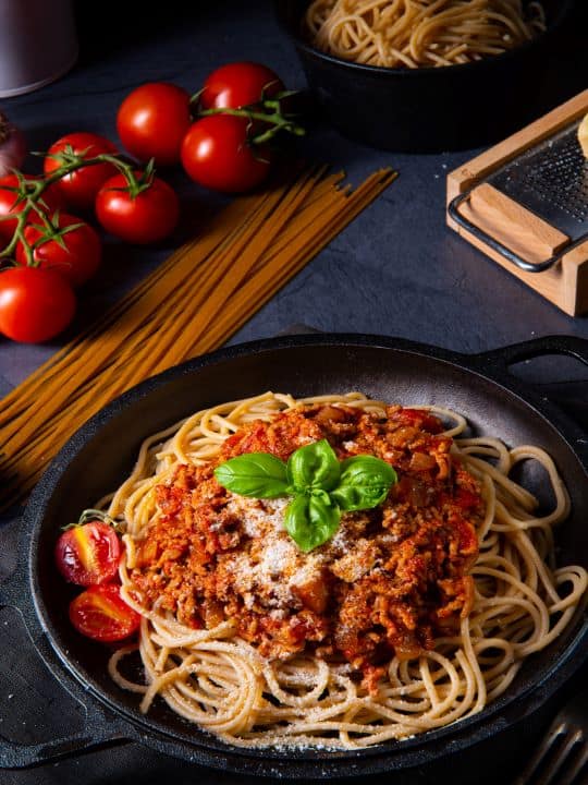 A black cooker contains cooked spaghetti with a kid-friendly pasta sauce on top. Some parmesan cheese is sprinkled over the pasta and green basil is in the middle of the cooker.