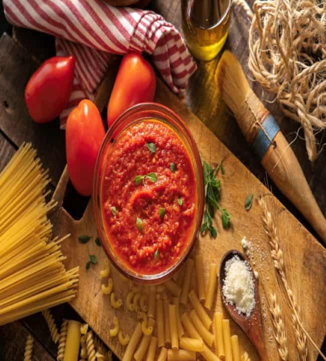 A bowl of red pasta sauce rests on a cutting board. It is surrounded by uncooked spaghetti, three tomatoes, a spoonful of parmesan, and some utensils.