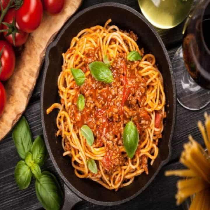 A black plate contains cooked spaghetti with red pasta sauce. The spaghetti has green basil leaves on top. Green basil leaves are near the plate with uncooked pasta on the other side.