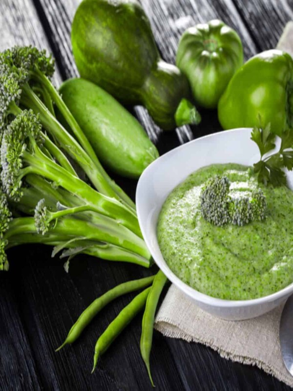 Some green veggies sit on a wooden table. They are placed near a bowl of a magical green sauce.