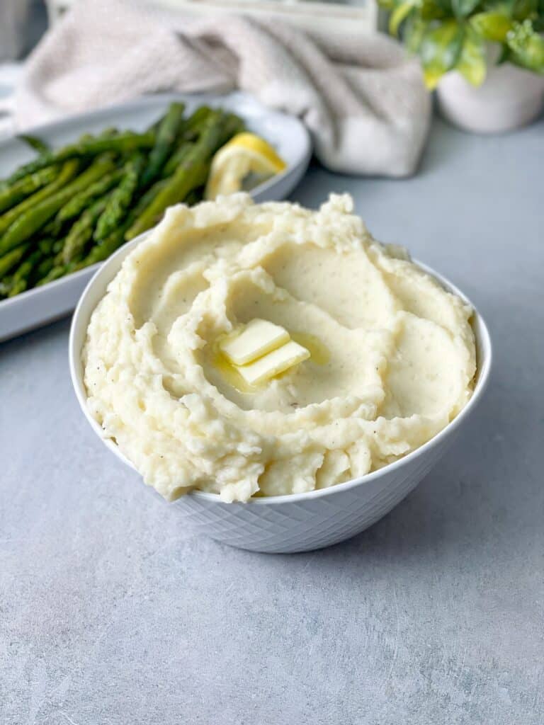 a bowl of creamy mashed potatoes served with pieces of butter on top alongside boiled asparagus