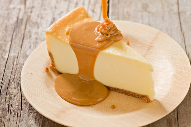A slice of New York-style cheesecake sitting on a bamboo plate and a Dulce de Leche poured on top.