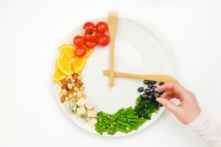 Colorful food and cutlery arranged in the form of a clock on a plate. Olive in hand. Intermittent fasting, diet, weight loss, lunch time concept.