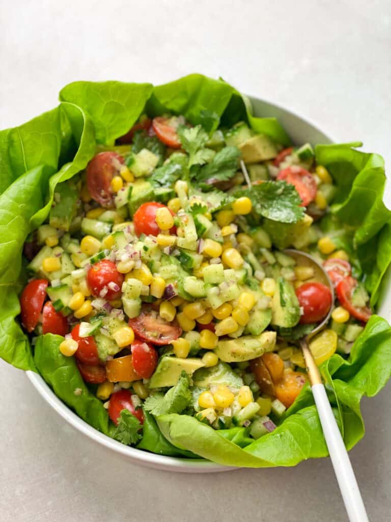 a bowl of avocado salad with cherry tomatoes, corn, cucumbers, and chopped onions drizzled with lime cilantro dressing.