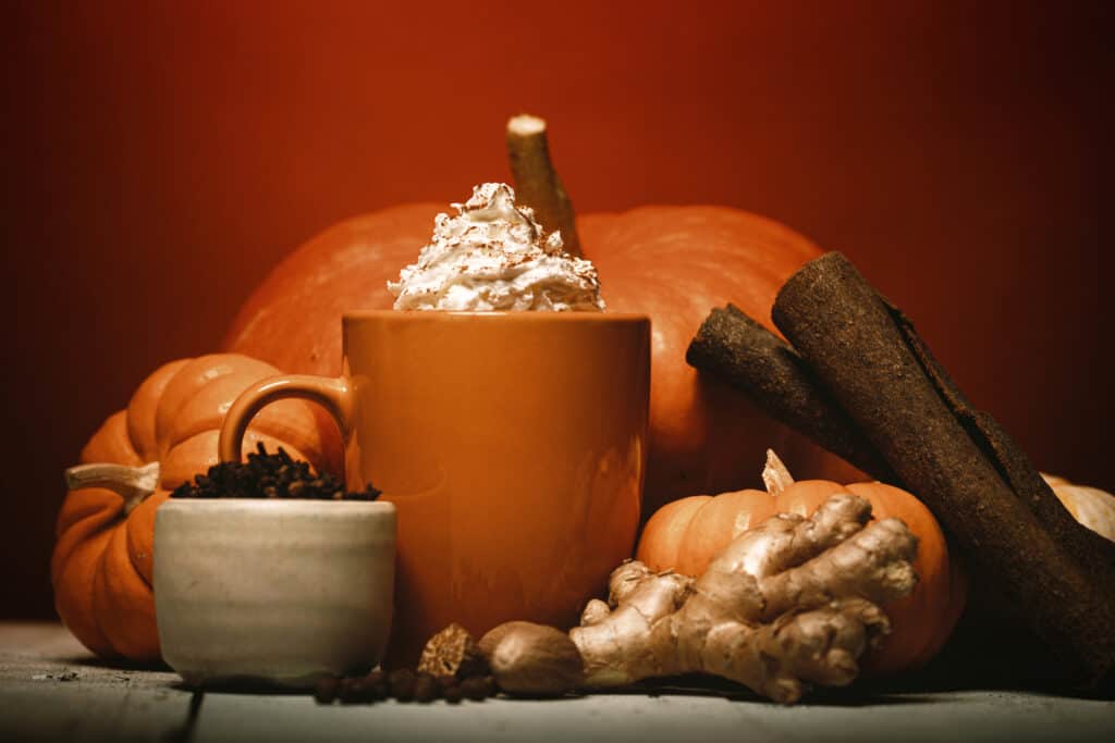 An Autumn favorite hot drink, the pumpkin spice flavored coffee, tea, or chai, complete with fresh pumpkins and whipped cream topping.  Cinnamon sticks, ginger, cloves, and allspice surround the mug of spicy warm goodness.  Rustic wood table.