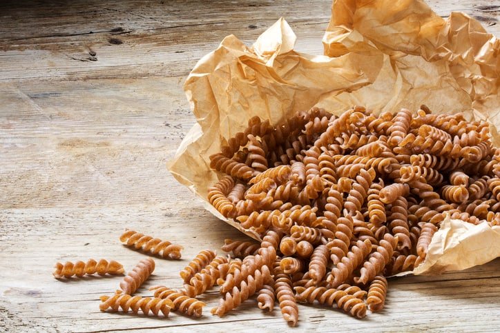 whole meal pasta from organic whole grain spelt falling from a paper bag on a rustic wooden table.