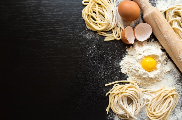 Raw homemade pasta with flour nest, egg in its center and a wood rolling pin on black background.