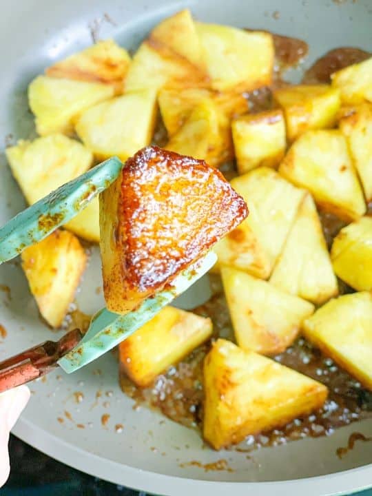A pan contains sliced pineapple with brown caramel and butter around them. Above the pan, tongs hold one fried pineapple slice.