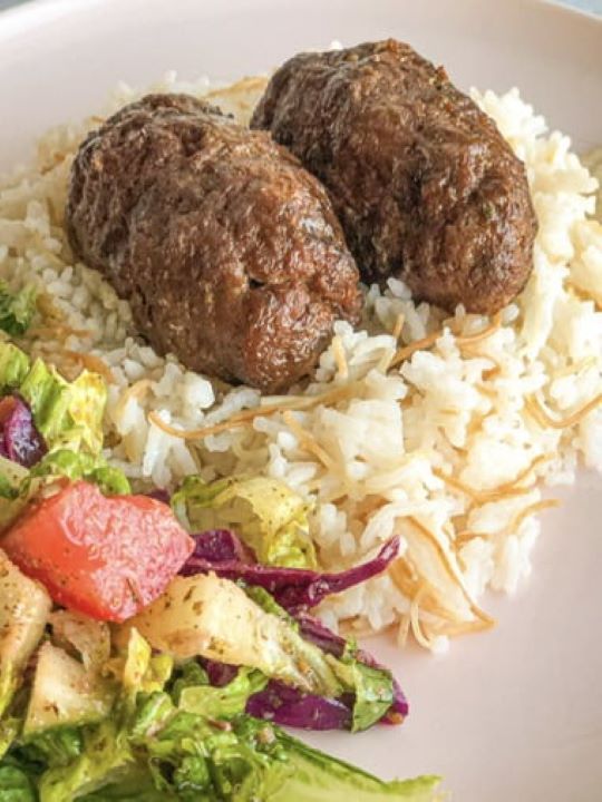 The grilled kofta kebabs are placed on the top of the plate. Beneath them, there is cooked rice, and near it vegetable salad lines.