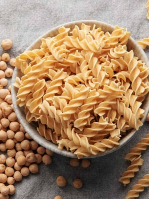 Against a grey background, some healthy chickpea grains are scattered near a bowl. The bowl contains uncooked pasta.