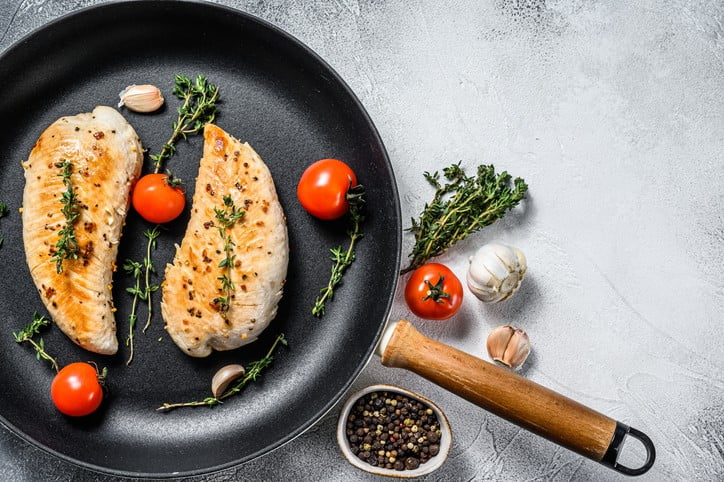 tender juicy cooked chicken breast fillet in a frying pan topped with rosemary alongside fresh tomatoes, garlic cloves, and pepper