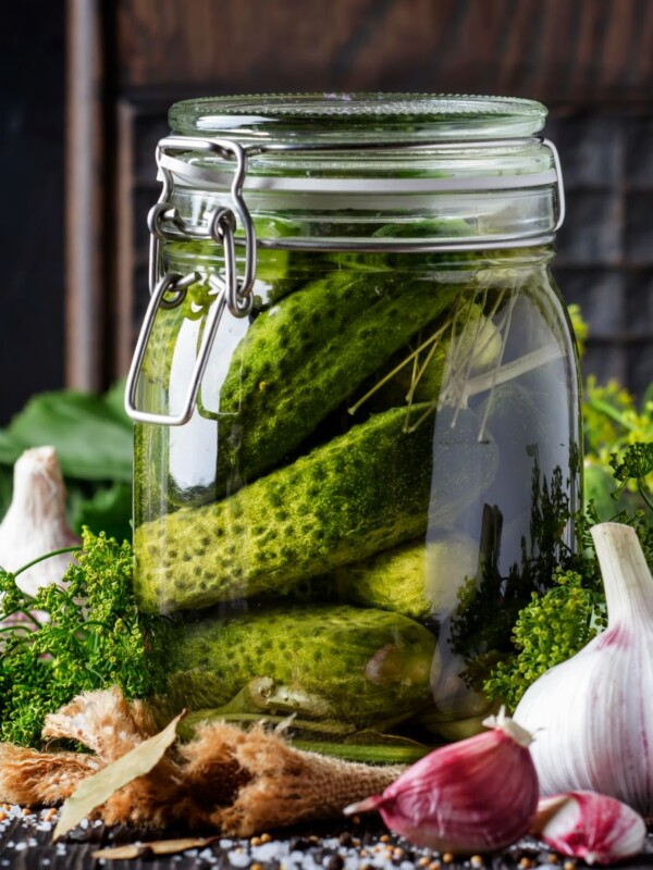 Big jar of cucumber pickles next to herbs and garlic cloves.