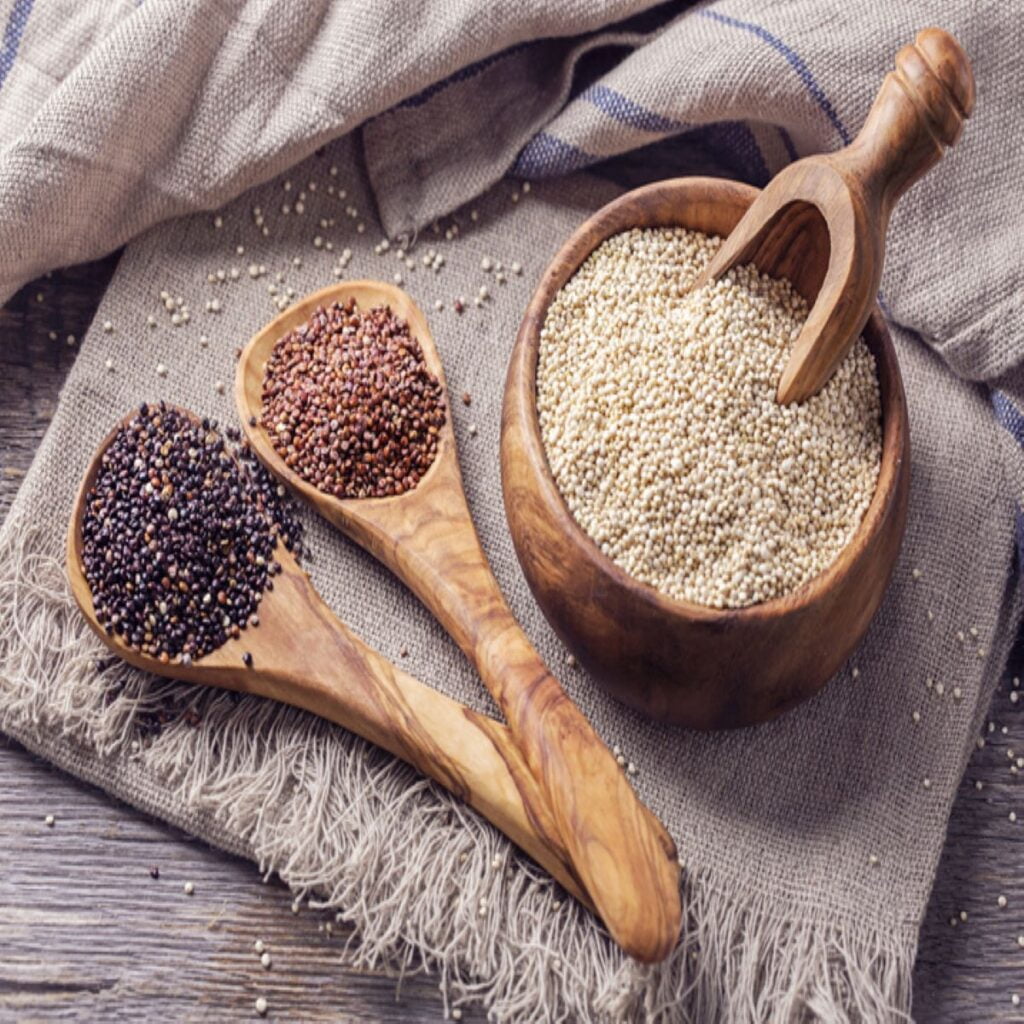 Burlap cloth is placed on a wooden background. On the cloth there are two wooden spoons and pots filled with quinoa of different colors as per rice vs quinoa.