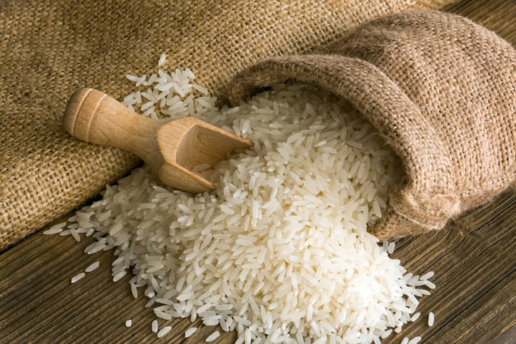 White rice seeds come out of a burlap sac. A wooden spoon scoops some of the rice which might not be healthier than quinoa against a wooden background.