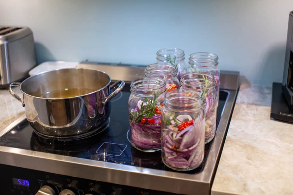 Homemade sweet and spicy pickled red onions sliced in a large mason glass jar, made with chili peppers, garlic, pepper corns, rosemary and apple cider vinegar sitting on a stove top.
