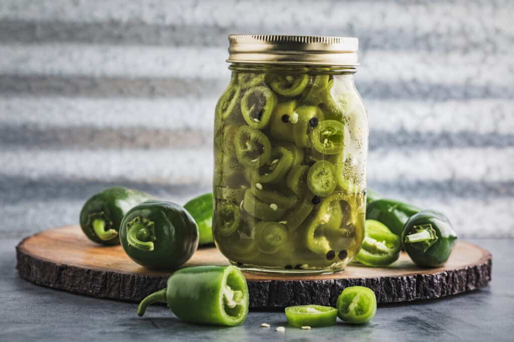 Jar of  quick pickled jalapeno peppers surrounded by fresh green jalapeno peppers 
