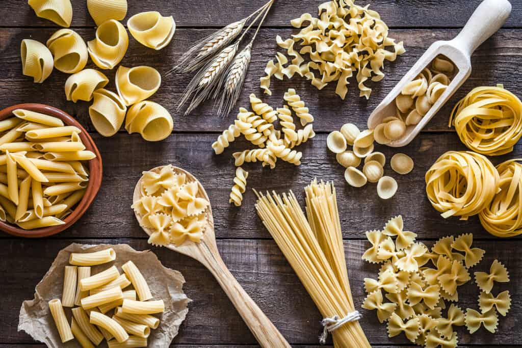 Top view of a rustic wooden table filled with a large Italian pasta variety. The types of pasta included are spaghetti, orecchiette, conchiglie, rigatoni, fusilli, penne and tagliatelle. 