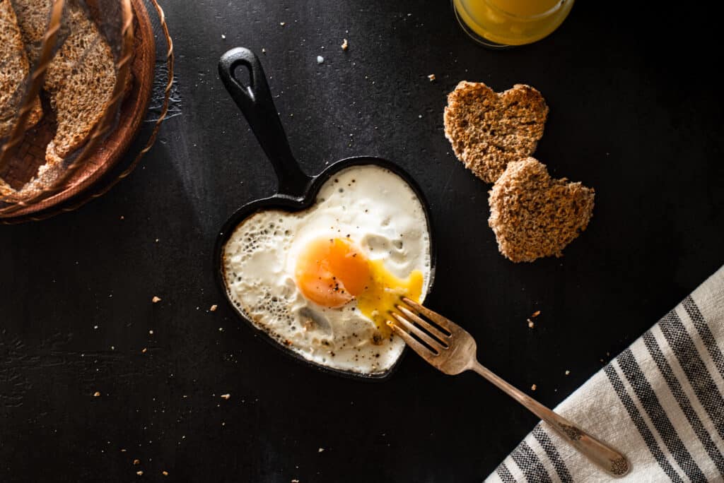 Flat lay of sunny side up egg in egg shaped cast iron skillet, orange juice, yellow berries and heart shaped bread