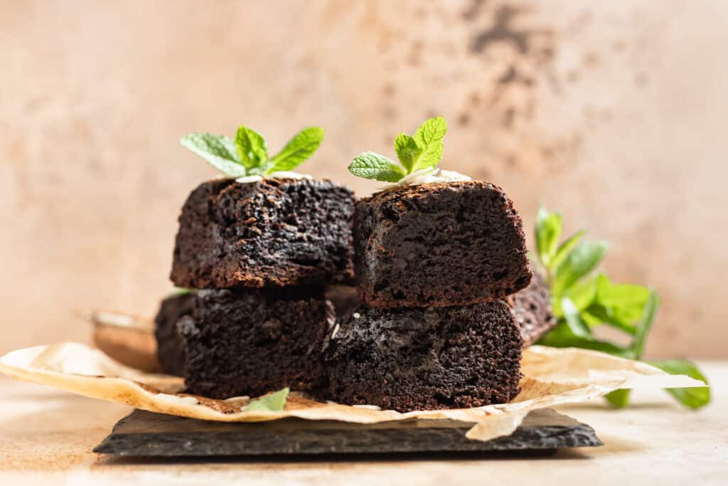 Stack of brownies made of dark chocolate served with cocoa powder and mint on baking paper, brown background. Tasty fudge and chewy dessert with melting pieces of chocolate.