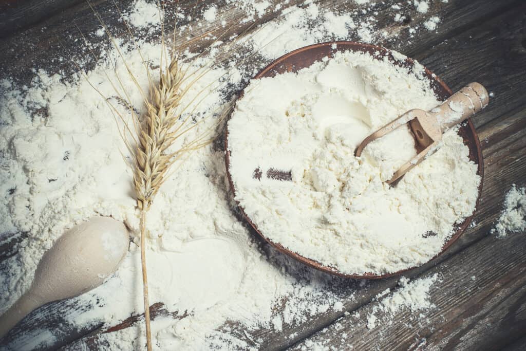 White flour and spikelets of wheat for baking bread lies on a wooden brown surface.