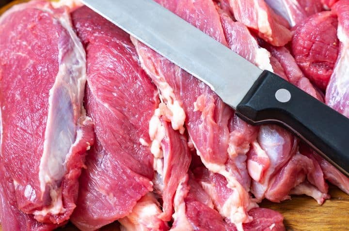Large chunks of beef are place on a cutting board with a big and sharp knife on top of them.