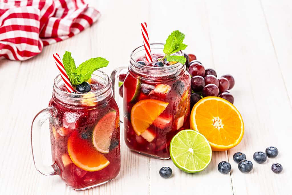 Two mason jars with a handle that contain a refreshing drink red drink with chunks of fruits.