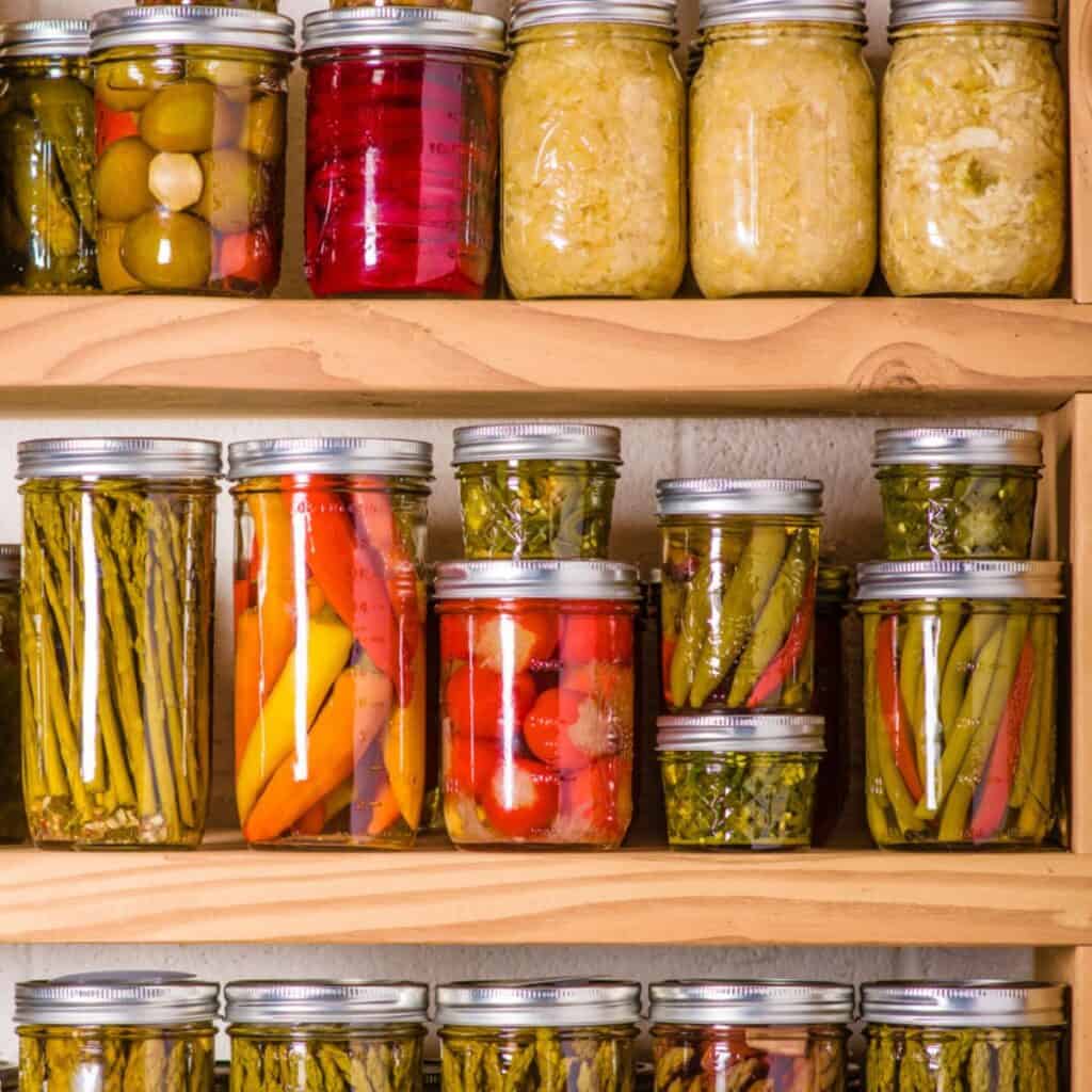 A stack of mason jars in different sizes that contain pickled veggies, such as carrots, asparagus, and reddish.