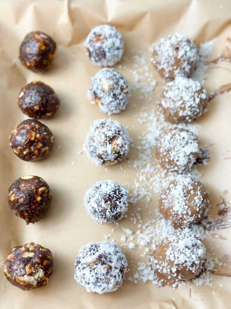 Date balls placed on a parchment paper after being frozen. Some are covered with coconut only, some are covered with chocolate and coconut and others are not covered with anything. 