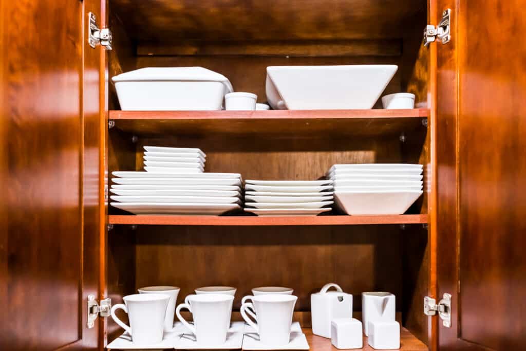 Open wooden kitchen cabinet door cupboard with many white dishes, plates, cups on shelves closeup